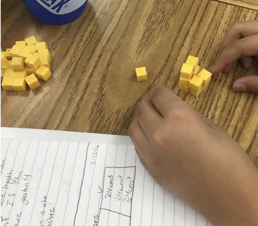 A student's hands are shown building a rectangular prism with yellow plastic cubes.